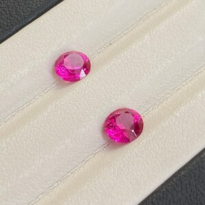 May include: Two round, pink gemstones on a white background. The gemstones are set in a tray with a black border.