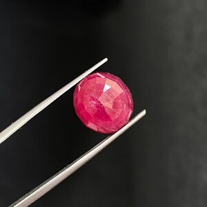 May include: A bright red ruby gemstone with a faceted cut, held by tweezers against a black background.