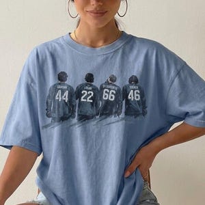 May include: A light blue t-shirt with a graphic of four figures wearing jerseys with the numbers 44, 22, 66, and 46. The names Graham, Logan, D Laurenty, and Tucker are above the numbers. The text "Blue Jean" is at the bottom.