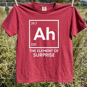 May include: A maroon t-shirt with a white periodic table-style graphic that reads "Ah" with the numbers 29.7 and 230, and the text "THE ELEMENT OF SURPRISE". The shirt is hanging on a clothesline.