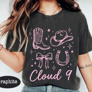 May include: Graphite-colored t-shirt featuring a pink cowboy boot, hat, bow, horseshoe, and the words "Cloud 9." The design includes stars and sparkles, creating a playful, Western-themed graphic.