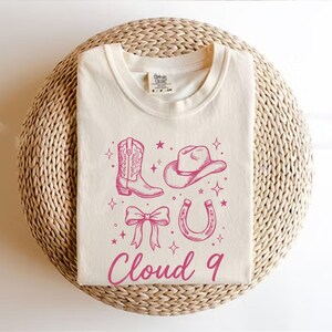 May include: Cream-colored t-shirt featuring a pink cowboy boot, hat, bow, and horseshoe graphic with stars. The text "Cloud 9" is printed in pink below the graphics. The shirt is displayed on a woven, circular surface.