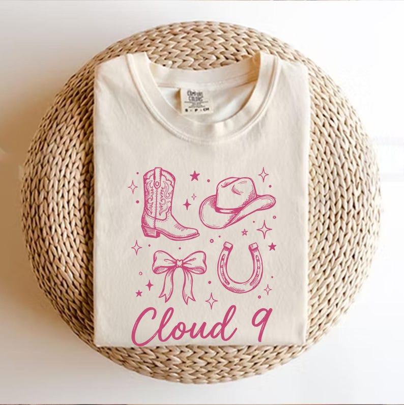 May include: Cream-colored t-shirt featuring a pink cowboy boot, hat, bow, and horseshoe graphic with stars. The text "Cloud 9" is printed in pink below the graphics. The shirt is displayed on a woven, circular surface.