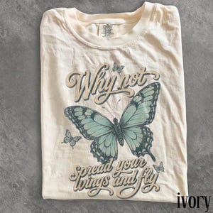 May include: An ivory t-shirt featuring a large, detailed teal butterfly graphic in the center. The text "Why not" appears above the butterfly, and "spread your wings and fly" is printed below in a cursive font. Smaller butterflies surround the design.