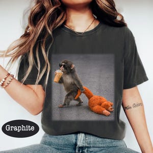 May include: Graphite-colored t-shirt featuring a graphic of a monkey wearing sunglasses and holding an iced coffee, dragging an orange stuffed animal. The shirt is a comfortable, casual style.