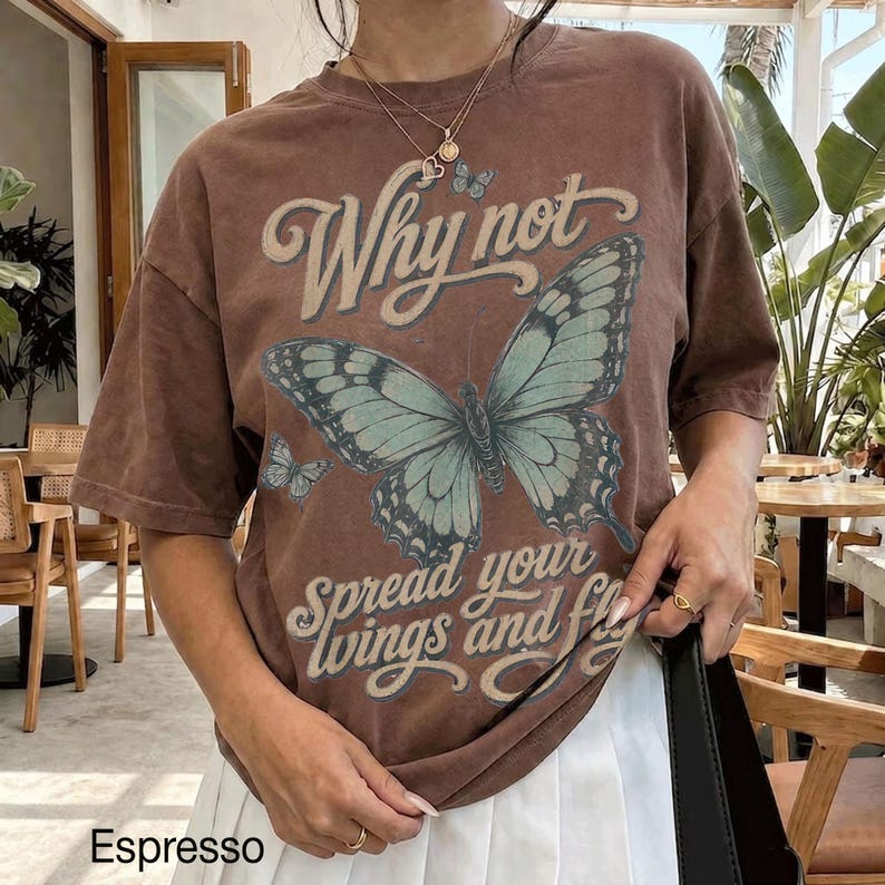 May include: A brown short-sleeve t-shirt featuring a large light blue butterfly graphic in the center. The text "Why not" appears above the butterfly, and "Spread your wings and fly" is printed below it. A small butterfly is also visible.