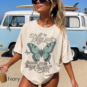 May include: An ivory short-sleeve t-shirt featuring a large teal butterfly graphic in the center. The text "Why not" appears above the butterfly, and "spread your wings and fly" is printed below it in a decorative script font.