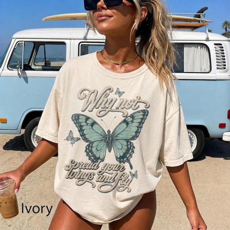 May include: An ivory short-sleeve t-shirt featuring a large teal butterfly graphic in the center. The text "Why not" appears above the butterfly, and "spread your wings and fly" is printed below it in a decorative script font.