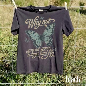 May include: A charcoal gray short-sleeve t-shirt featuring a large teal butterfly graphic in the center. The text Why not spread your wings and fly is printed in a light tan script font above and below the butterfly, with two small butterflies.