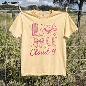 May include: Butter-colored t-shirt with a pink graphic of a cowboy boot, hat, bow, and horseshoe, with the text "Cloud 9" in a script font. The shirt is hanging on a clothesline outdoors.