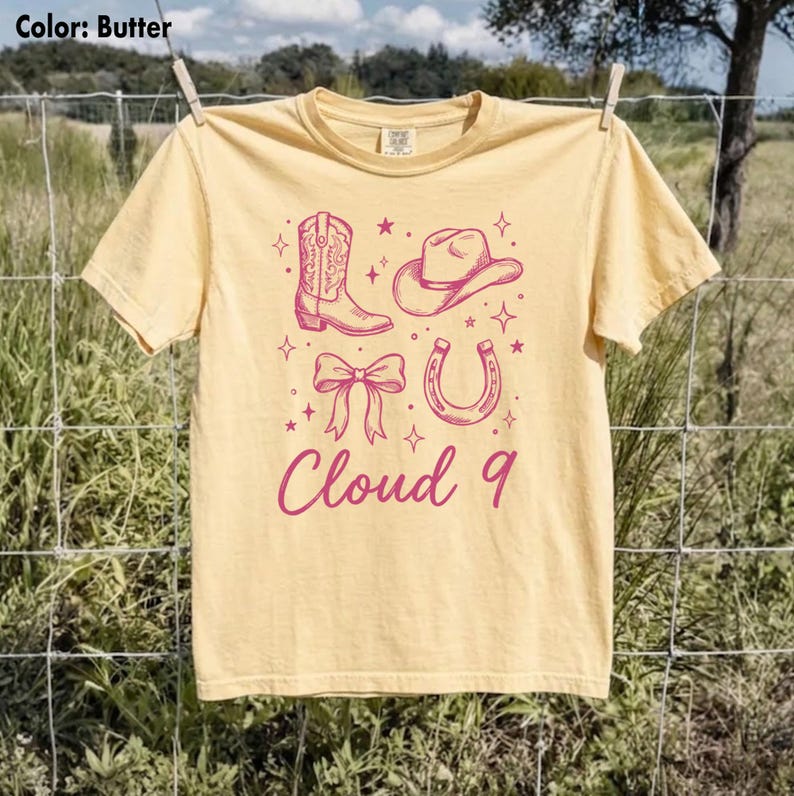 May include: Butter-colored t-shirt with a pink graphic of a cowboy boot, hat, bow, and horseshoe, with the text "Cloud 9" in a script font. The shirt is hanging on a clothesline outdoors.