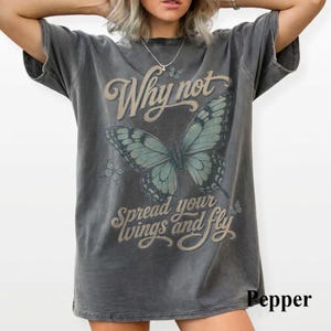 May include: A charcoal gray short-sleeve t-shirt featuring a large teal butterfly graphic in the center. The text Why not spread your wings and fly is printed in a tan script font. The shirt has a distressed, washed-out appearance throughout.