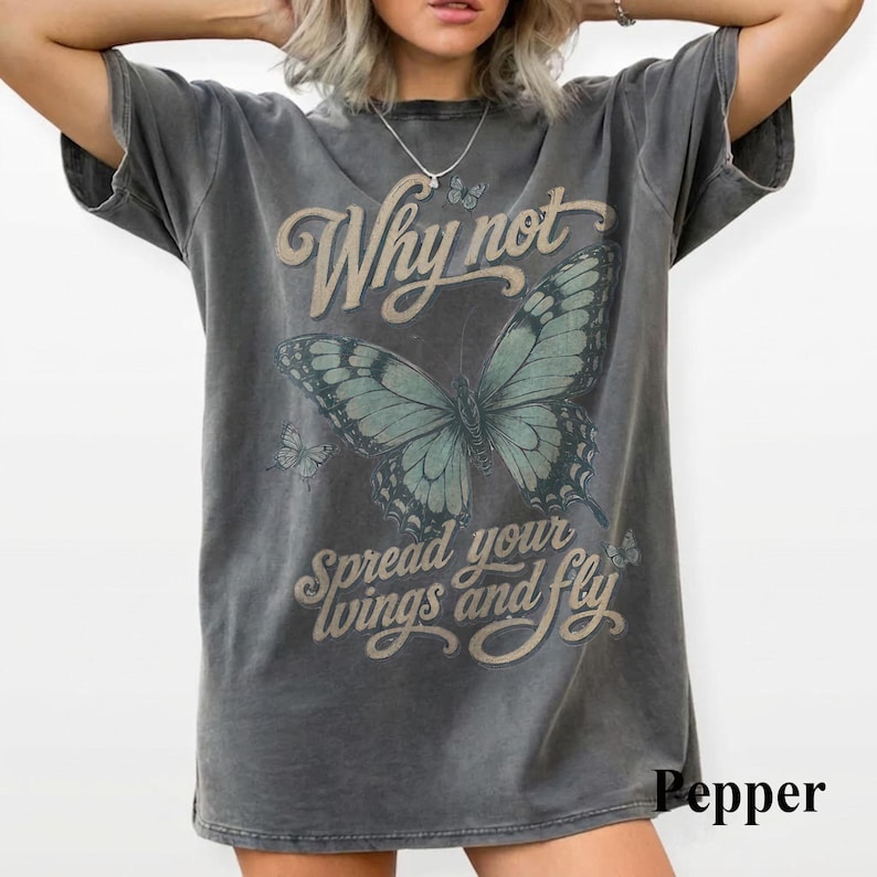 May include: A charcoal gray short-sleeve t-shirt featuring a large teal butterfly graphic in the center. The text Why not spread your wings and fly is printed in a tan script font. The shirt has a distressed, washed-out appearance throughout.
