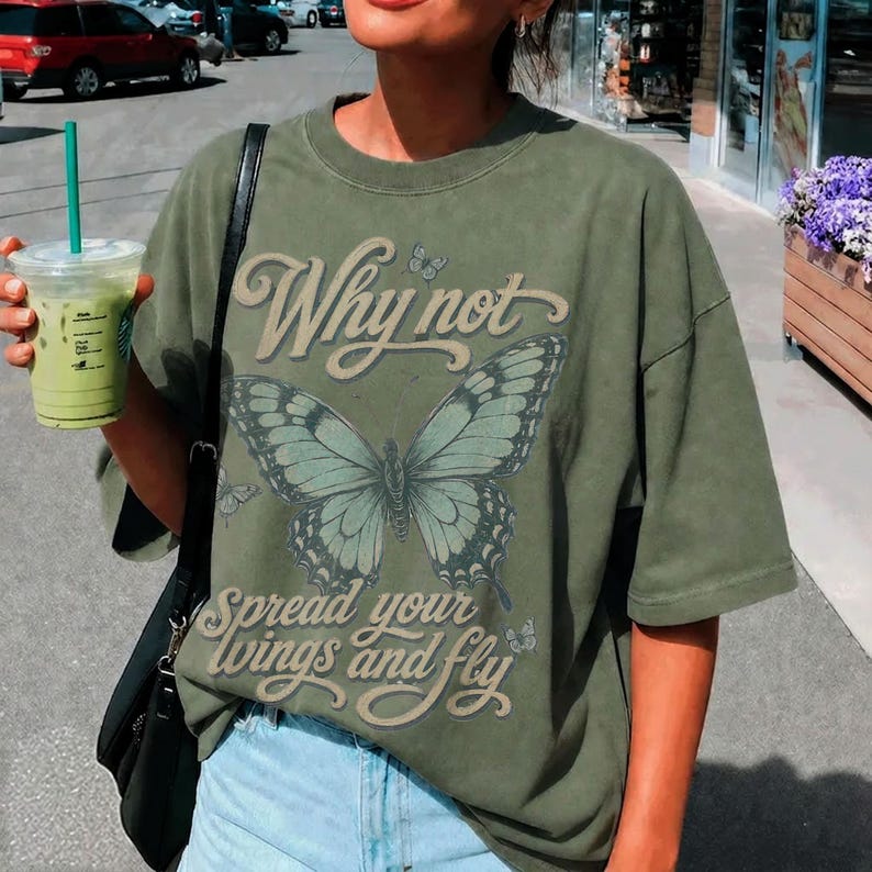 May include: An olive green short-sleeve t-shirt featuring a large butterfly graphic in the center. The text "Why not" is printed above the butterfly, and "spread your wings and fly" is printed below in a cursive font. The shirt has a crew neckline.