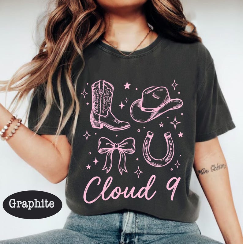 May include: Graphite-colored t-shirt featuring a pink cowboy boot, hat, bow, horseshoe, and the words "Cloud 9." The design includes stars and sparkles, creating a playful, Western-themed graphic.