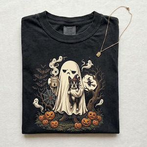 May include: A black crew neck t-shirt featuring a graphic of a ghost holding a mug labeled Hocus Pocus and a Boston Terrier wearing a witch hat. The design includes orange pumpkins, flying bats, a spider web, and small ghosts. A thin gold necklace is included.