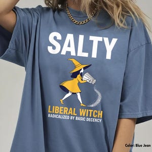 May include: Blue jean-colored t-shirt with the word "SALTY" in white block letters. Below, a cartoon witch in a yellow dress and hat pours salt from a shaker. The text "LIBERAL WITCH RADICALIZED BY BASIC DECENCY" is below the witch.