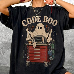 May include: A black short-sleeve t-shirt featuring a graphic print of a ghost standing behind a red medical cart. The text CODE BOO is printed above the ghost. The cart includes a monitor with a heart rhythm, a stethoscope, and an oxygen tank.