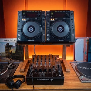 以下が含まれることがあります： 木製スタンドに設置された2台の黒いPioneer CDJ-850デジタルメディアプレーヤーと、その下の黒いPioneer DJミキサー。左側には黒いヘッドホンがあり、木製のデスク上の機器の両脇にはレコードが見える。