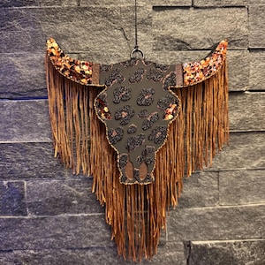 May include: A hanging wall ornament shaped like a bull skull, featuring a black base with a glittery leopard print pattern. The horns are adorned with copper-colored sequins, and long, thin, metallic copper-colored fringe hangs from the horns and base.