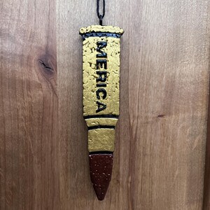 May include: A gold-painted bullet-shaped ornament with the word "MERICA" written vertically on the side. The ornament is hanging from a black string.