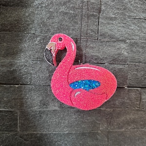 May include: A pink, glittery flamingo-shaped ornament. The flamingo has a black and gold beak, a blue glitter accent, and a pink glitter body. The ornament is suspended against a dark grey brick wall.