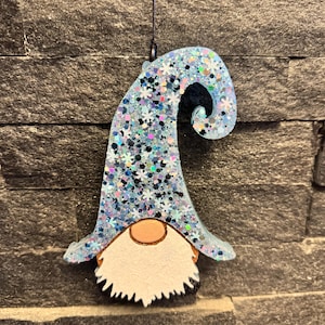 May include: A hanging gnome ornament featuring a tall, curved hat filled with blue, silver, and iridescent glitter mixed with small white snowflake shapes. The gnome has a round peach-colored nose and a textured white beard hanging from the hat.