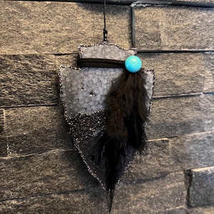 May include: A hanging arrowhead-shaped ornament featuring a textured gray surface with a dark glittery base. It is accented with a black suede cord, a single bright turquoise bead, and a cluster of soft black feathers hanging from the side.