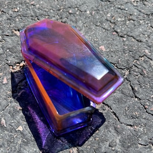 May include: A small, iridescent coffin-shaped box with a hinged lid. The box is made of glass and has a purple, blue, and orange colour scheme.