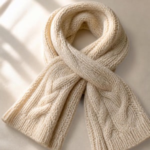 May include: A cream-colored cable knit scarf featuring a thick, chunky texture with vertical braided patterns running along the length of the fabric. The scarf is arranged in a loose loop, showcasing the detailed knit construction and ribbed edges.