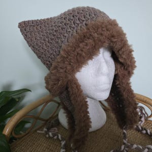 May include: A crocheted hat with a pointed top and ear flaps. The hat is a muted brown color with a fluffy, faux fur trim in a darker brown. The ear flaps have braided ties with white accents. The hat is displayed on a white mannequin head.