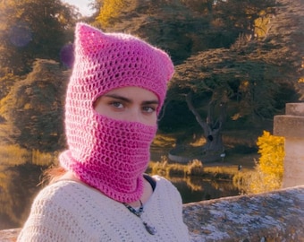 May include: A person wearing a pink crocheted balaclava with cat ears. The person is wearing a white sweater and is standing in front of a stone wall.