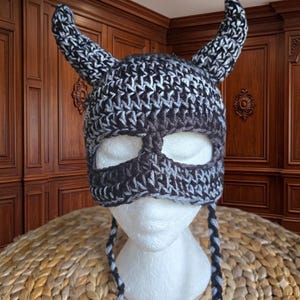 May include: A handmade crochet hat in a Viking helmet style. The hat features a dark gray and white color scheme, with two curved horns and a face mask with eye openings. Braided ties hang from the sides. The hat is displayed on a white mannequin head.