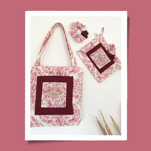 Könnte beinhalten: Ein Set aus Tragetasche, Scrunchie und Topflappen. Die Artikel weisen ein weißes und rosa Blumenmuster mit einem bordeauxfarbenen Quadrat auf. Jeder Artikel hat ein kleines Etikett mit Text. Das Set wird auf weißem Hintergrund präsentiert.