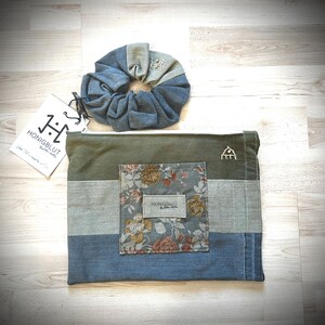 Könnte beinhalten: Eine rechteckige Reißverschlusstasche mit horizontalen Farbblöcken in Olivgrün, Hellgrau und blauem Denim, einer mittigen Tasche mit Blumenmuster und einem passenden Haargummi aus blauem Denim. Ein weißes Etikett zeigt den Text HONIGBLUT.
