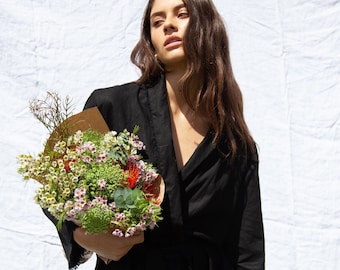 May include: A woman wearing a black linen dress with a frayed hem, holding a bouquet of flowers wrapped in brown paper. The flowers include white, pink, and orange blooms.