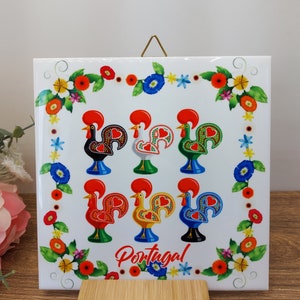 May include: White ceramic tile with a decorative border of colorful flowers and leaves. Six colorful rooster illustrations are arranged in a grid pattern. The word "Portugal" is printed in the center of the tile.