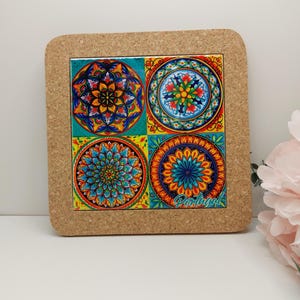 Pode incluir: Um descanso de panela com suporte de cortiça com um azulejo quadrado de cerâmica que apresenta quatro desenhos circulares coloridos com padrões intrincados. O azulejo tem a palavra "Portugal" impressa nele.