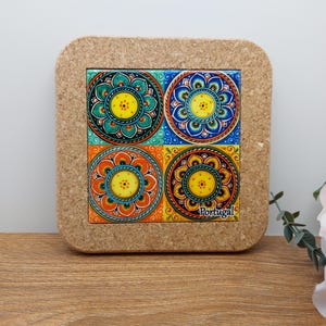Pode incluir: Um descanso de copo com suporte de cortiça com um azulejo quadrado de cerâmica com quatro desenhos circulares coloridos. O azulejo tem um esquema de cores amarelo, azul, verde e laranja. O texto "Portugal" está impresso no azulejo.
