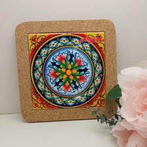 Pode incluir: Um descanso de copo com suporte de cortiça com um design circular colorido que apresenta uma flor amarela cercada por motivos florais azuis, verdes e vermelhos. O design lembra a cerâmica italiana tradicional.