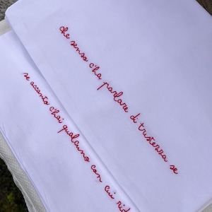 Può includere: Tessuto bianco con testo italiano ricamato in rosso. Il testo recita: "Che senso c'ha parlare di tristezza se accanto c'hai qualcuno con cui ridere."