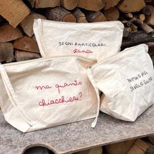 Può includere: Tre pochette con cerniera in tela bianco sporco con testo ricamato in rosso e nero. Le pochette sono disposte su una pila di legna da ardere. Il testo sulle pochette recita: "SEGNİ PARTICOLARI: stanca", "ma quanto chiacchieri?" e "PRIMA O POI SBAGLIO GIUSTO".