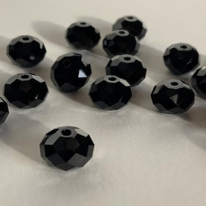 May include: A scattering of glossy, black faceted beads. Each bead is round with multiple flat surfaces, creating a sparkling effect. The beads are scattered on a light-coloured surface, ideal for jewellery making.