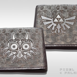 Könnte beinhalten: Zwei braune Ledergeldbörsen mit geprägten Designs. Eine zeigt die Majora-Maske, die andere das Triforce-Symbol, beide aus der Legend of Zelda-Serie. Die Geldbörsen haben einen kreisförmigen Musterhintergrund und werden auf einer weißen Oberfläche präsentiert.