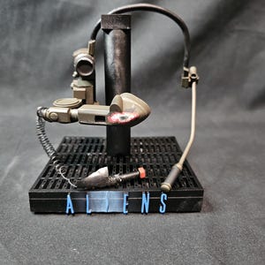 Może przedstawiać: Czarno-szara figurka akcji przedstawiająca broń science fiction z czerwonym światłem z boku. Figurka stoi na czarnej podstawie z nadrukiem słowa "ALIENS".