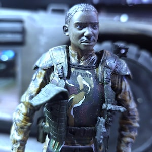 May include: A close-up of a military action figure wearing a camouflage uniform with a black vest and a tactical belt. The figure has a dark complexion and short black hair. The figure is holding a gun in its right hand.