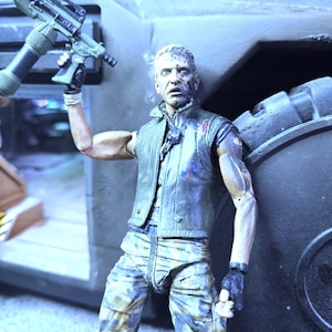 May include: A detailed action figure of a man with a distressed face, wearing a black vest, camouflage pants, and holding a black and grey gun. The figure is standing in front of a large, grey vehicle.