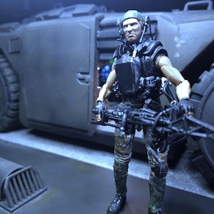 Pode incluir: Uma figura de ação militar com um padrão de camuflagem, segurando uma arma grande, em pé na frente de um veículo grande de cor cinza escuro.