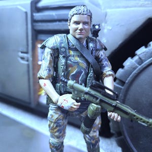 May include: A green and brown camouflage action figure of a soldier holding a gun. The figure is wearing a black harness and has a black and white patch on the chest that says "DIETRICH".