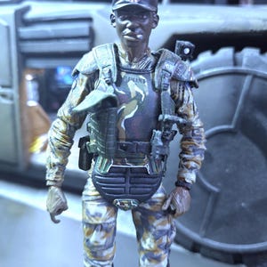 May include: A dark-skinned action figure wearing a camouflage uniform with a black vest and a blue, white, and red cap. The figure is holding a gun and has a serious expression on its face.